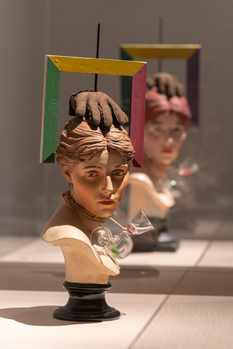 skulptur. kvindetorso med handkse på hovedet og ramme rundt om hovedet, penis på den ene kind