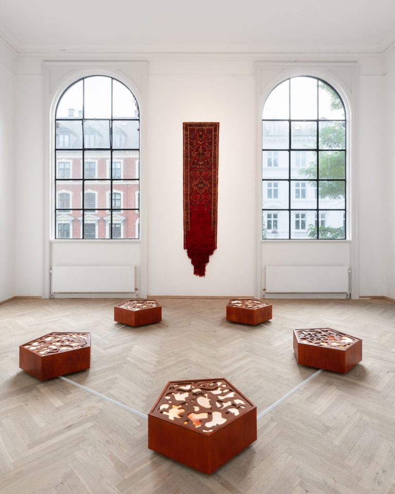 Et rødt tæppe hænger på væggen, fem træskulpturer står på gulvet i en pentagonform.