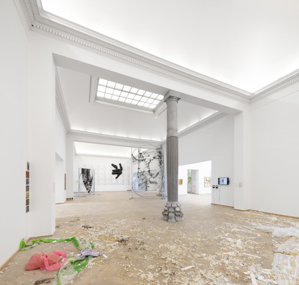 A large exhibition space with various artworks on the walls and floor. In the center of the room stands a column.