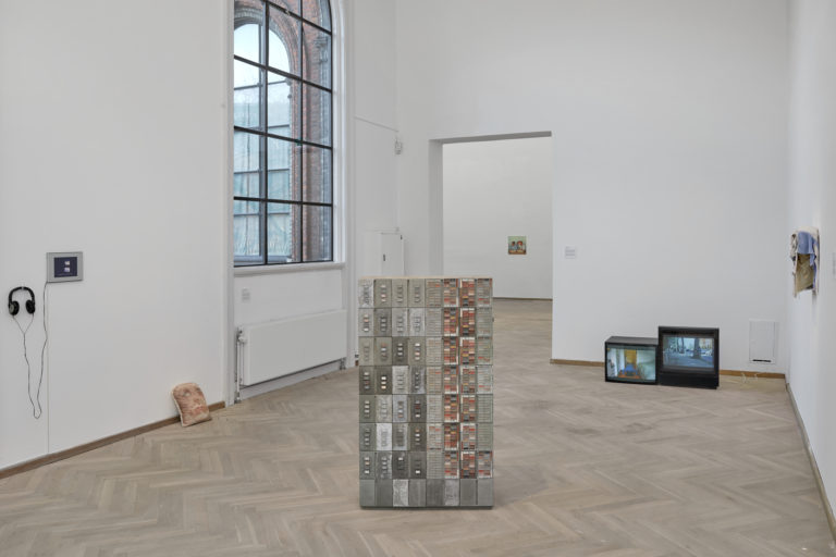 A bright exhibition room with a sculpture of concrete placed in the center of the room. Behind it, there are two old TV screens displaying video works. On each wall, there are hanging artworks in various media. Beneath the artwork hanging on the left-hand side, there is a cushion with a printed face on it.
