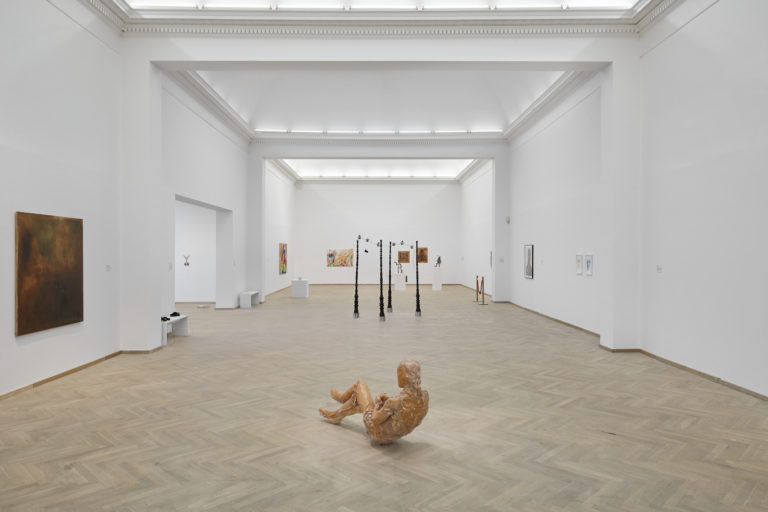 A bright exhibition room with several artworks placed around the gallery. In the foreground, there is a reclining human-like figure in a brownish color. Further back, there is a sculpture of four freestanding, dark, ornamented poles. Various works in different materials are hung on the walls.