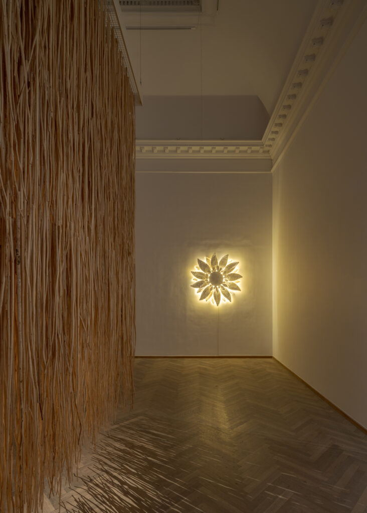 An exhibition room lit from the back wall by a hanging artwork in the shape of a flower, with light emerging from the flower’s petals. On the side of the room, a curtain made from the stalks of the jute plant is hanging.