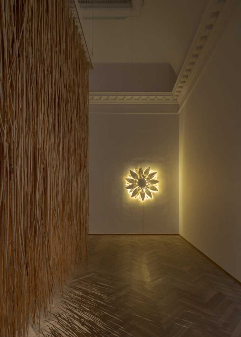 Et udstillingsrum belyst fra bagvæggen af et hængende værk i form af en blomst, hvor der bryder lys ud fra blomstens blade. På siden af rummet hænger der et gardin lavet af juteplantens stilke.