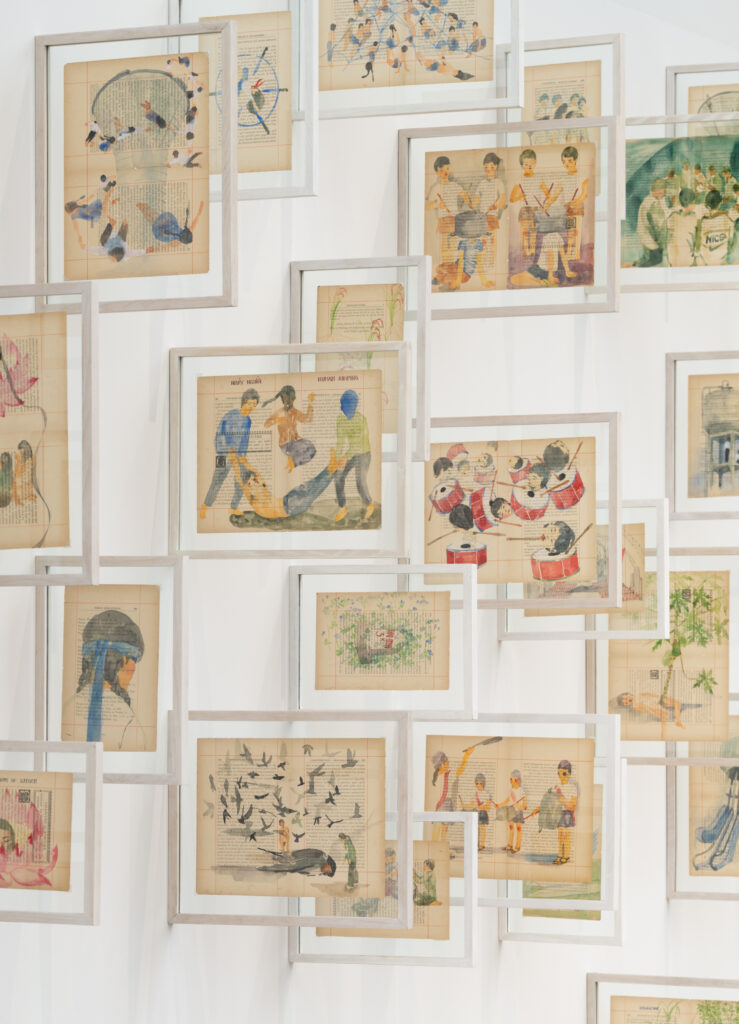 A bright exhibition room with many white picture frames hanging out from the wall. Each frame contains watercolor motifs painted on stretched silk.