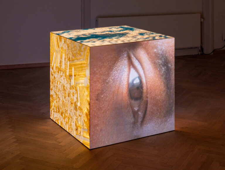 I et mørkt udstillingslokale, hvor en firkantet object står. På firkantens sider, projiceres der videoer med et øje og en guldbelagt lysekrone.
