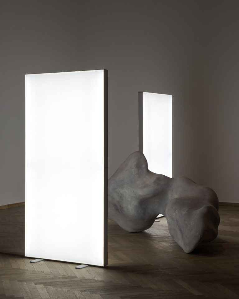 A dark exhibition room illuminated by a large standing white screen. Behind it is an organic concrete sculpture.