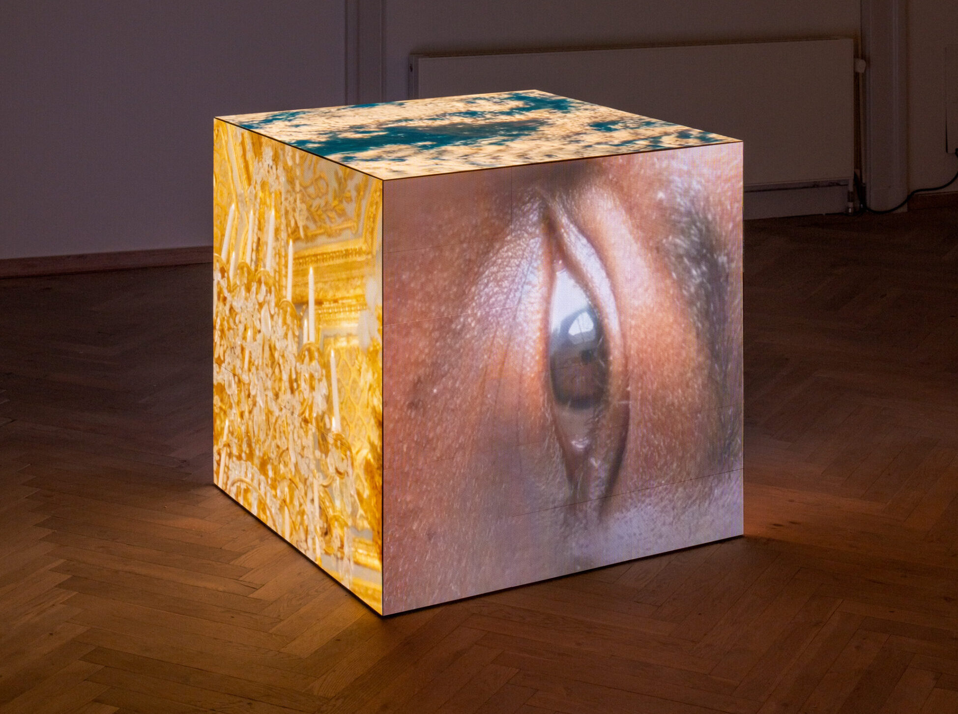 In a dark exhibition room, a square object stands. On the sides of the square, videos are projected showing an eye and a gold-plated chandelier.