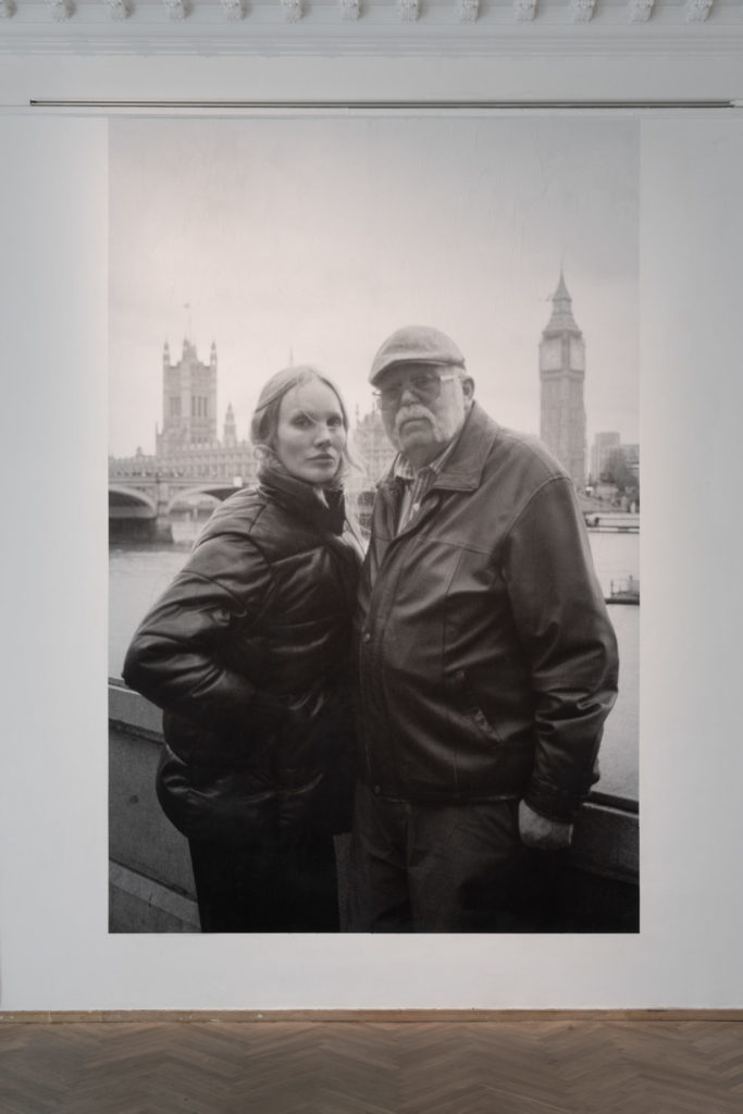 A photograph in an exhibition space, of two people standing next to each other.