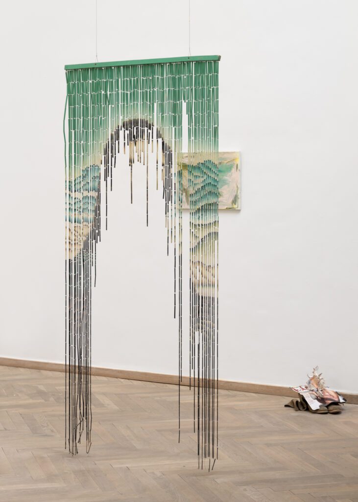 In a bright exhibition room, a door curtain hangs down from the ceiling. The curtain is split in the middle, and an image of a woman can be seen faintly.