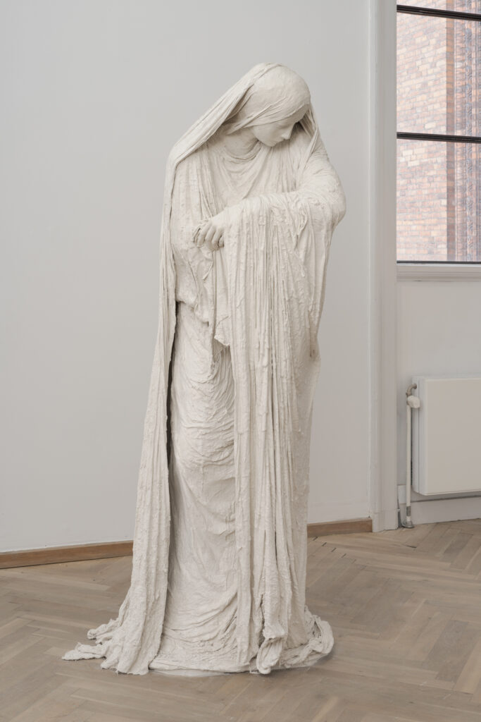 A sculpture of a woman dressed in garments covering her body and head. The woman is looking down at her arms, which are empty.