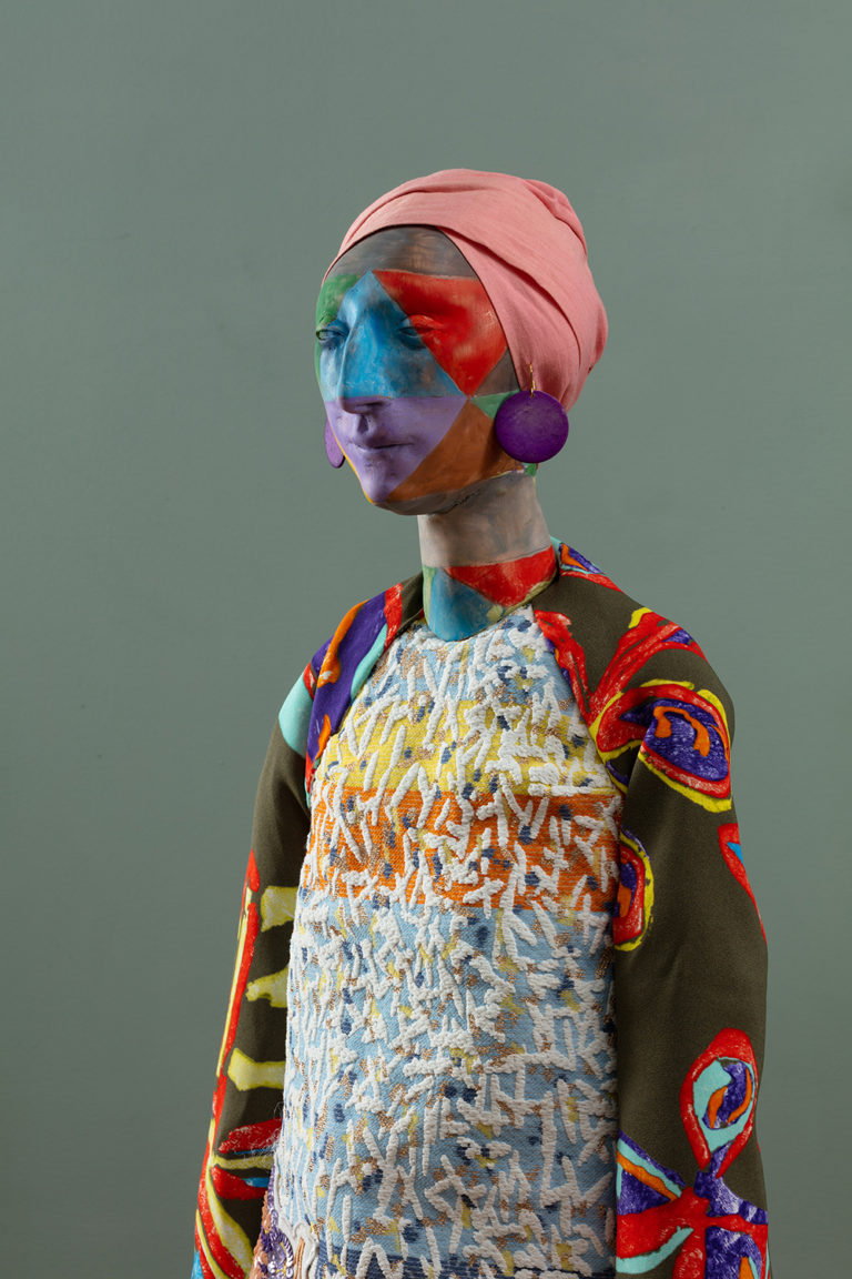 A sculpture of a human figure with a painted body. The figure is wearing a pink cloth on its head, and has purple earrings hanging from its ears. It is dressed in a colorful outfit.