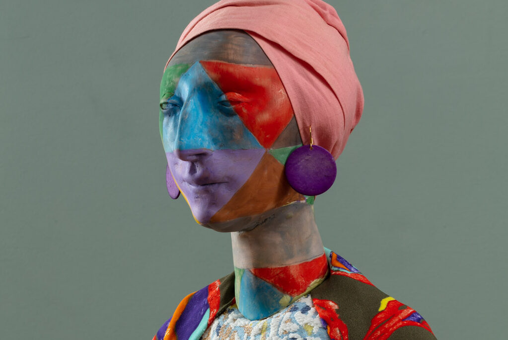 En skulptur af et menneske med malet krop. På hovedet bæres et lyserødt klæde og fra ørene hænger der lilla øreringe. Figuren er iklædt en farverig dragt.