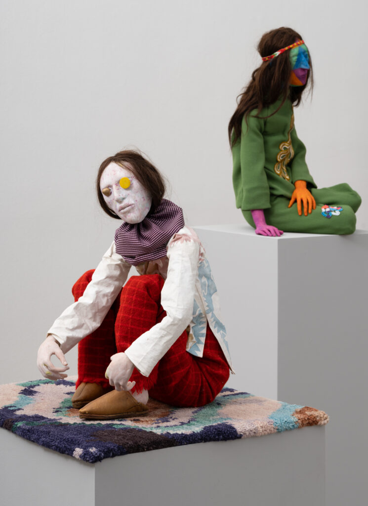 Two sculptures of human miniature figures placed on their own pedestals. Both with painted faces looking in different directions. The sculptures are dressed in various colorful clothing.