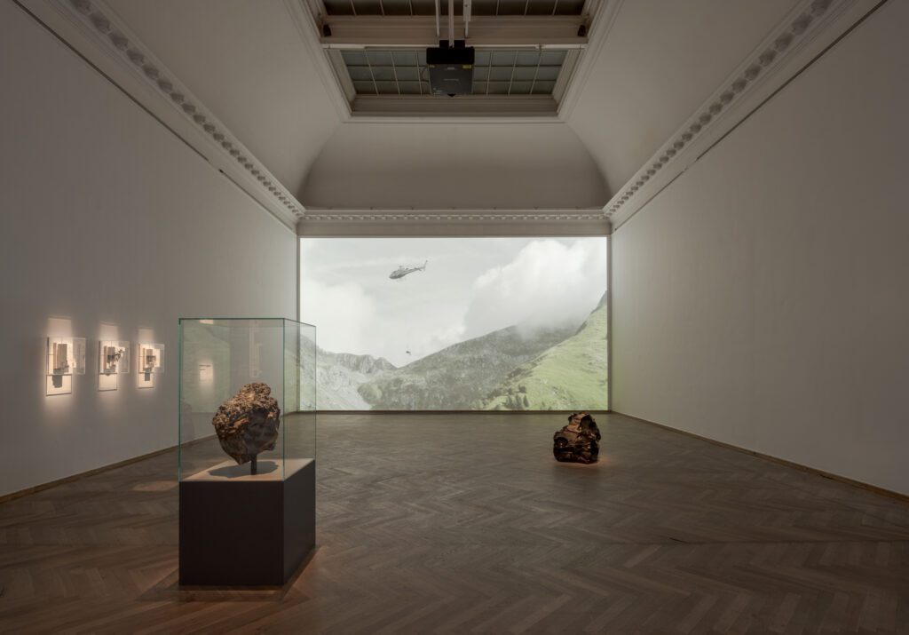 A dark exhibition room with two stone-like sculptures illuminated by spotlights, along with three white reliefs hanging on the wall. On the back wall of the room, there is a large video projection of a mountain landscape and a helicopter, from which a person is hanging by a rope.