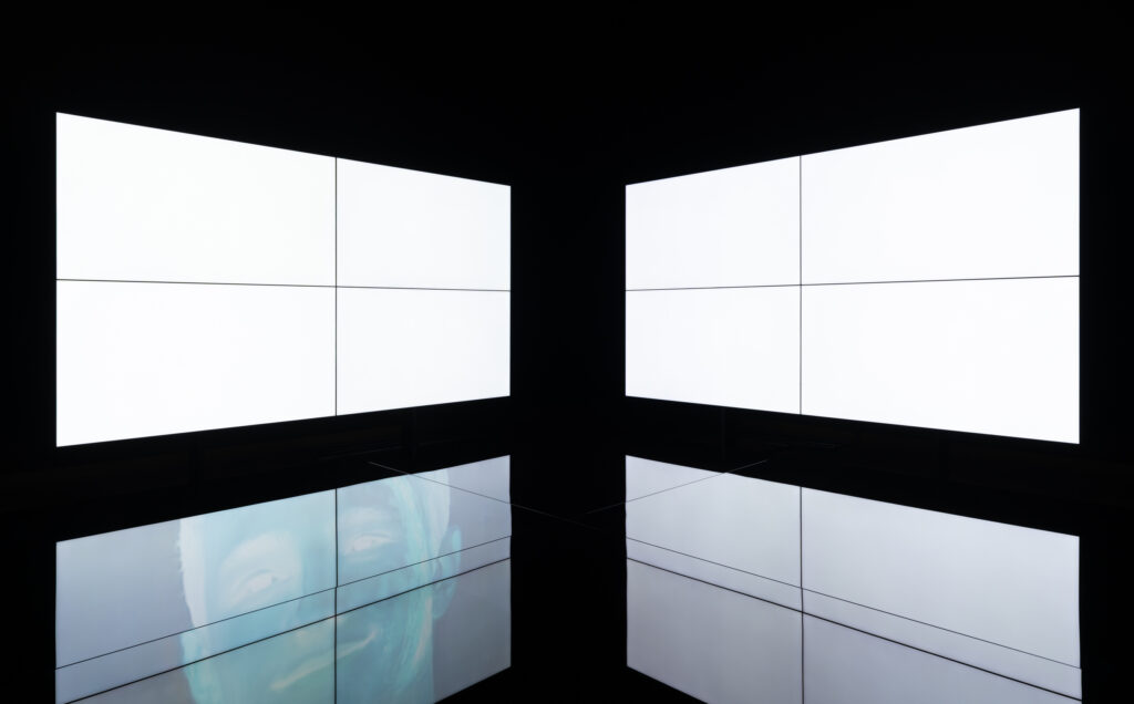 Two large screens illuminate a dark room. They are reflected in a black podium in the center. A bluish image is seen in the reflection.
