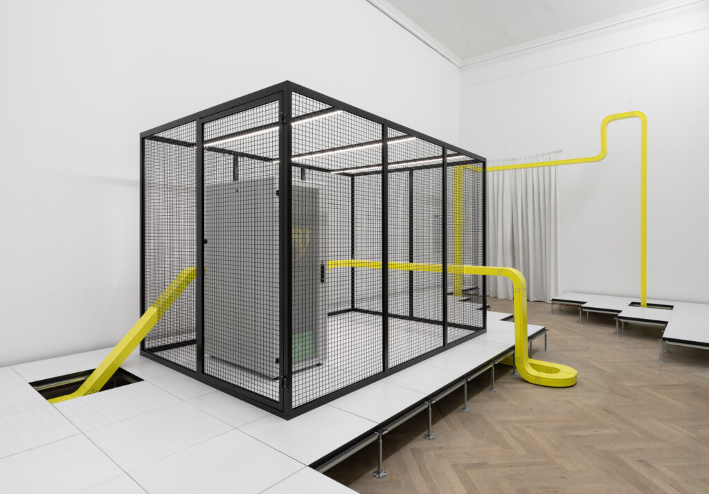 A bright exhibition room, where a black grid box stands on a podium. A yellow tube surrounds the box in the room.