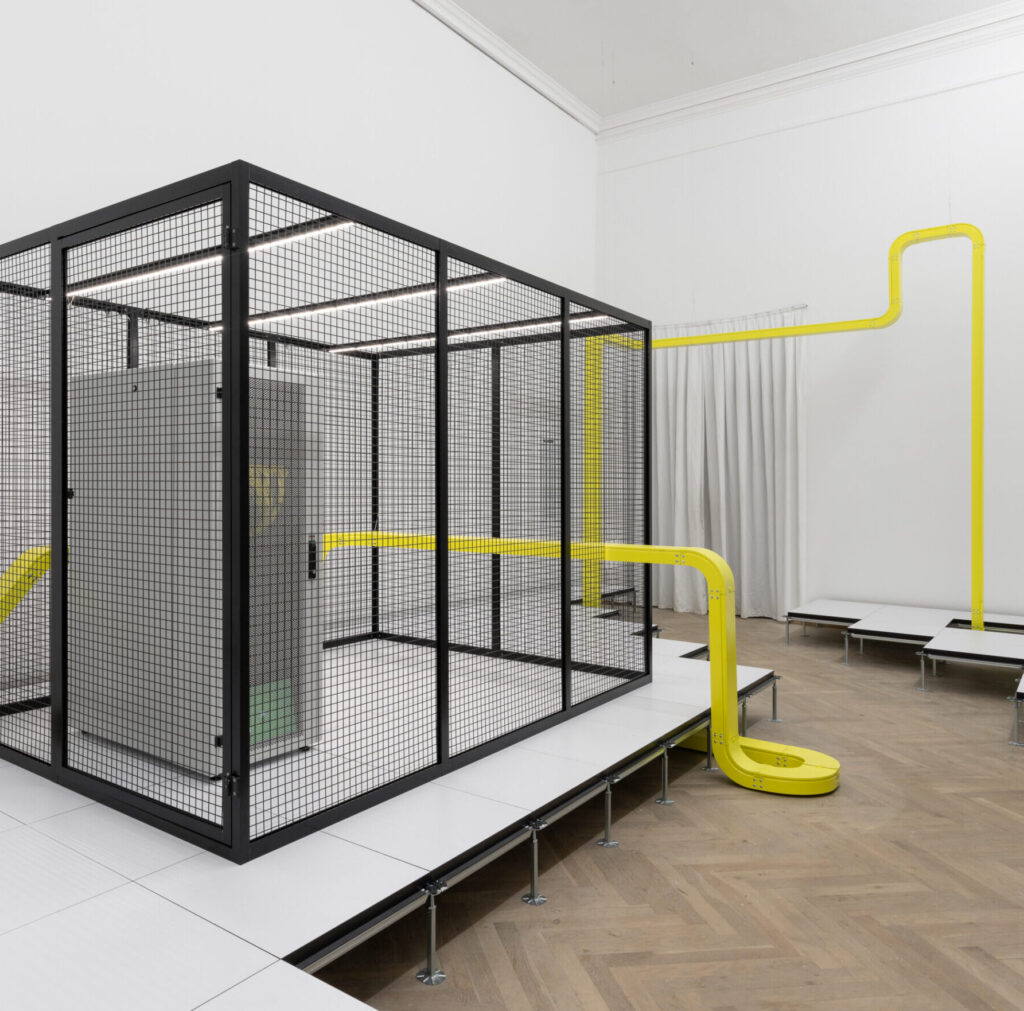 Et lyst udstillingsrum, hvor en sort gitterboks står på et podium. Et gult rør omringer boksen i rummet.