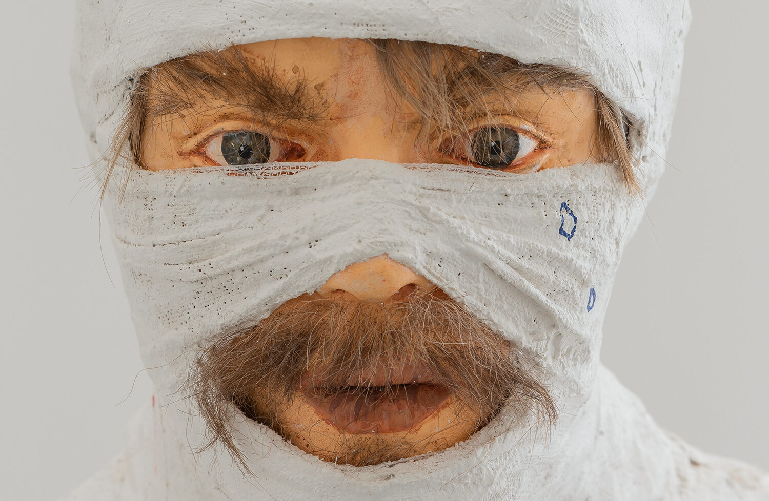 En skulptur af et mandligt ansigt med lys hud pakket ind i gips forbinding. Kun øjnene og munden er synlig. Det stikker brun ansigtsbehåring ud under gipsen.