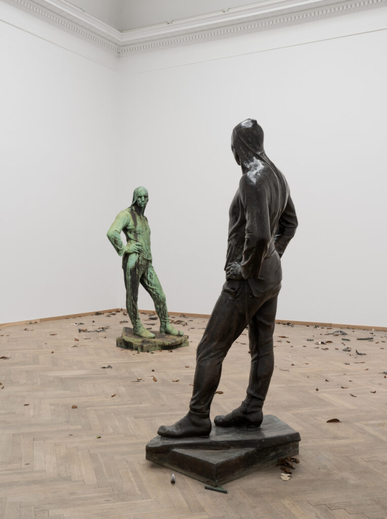 A bright exhibition room with two bronze sculptures, one more weathered than the other, standing opposite each other. The sculptures are almost identical, depicting a male human body draped in cloth. The figures are looking directly at each other, with one hand resting on their hips. Small leaves of bronze are spread out on the floor.
