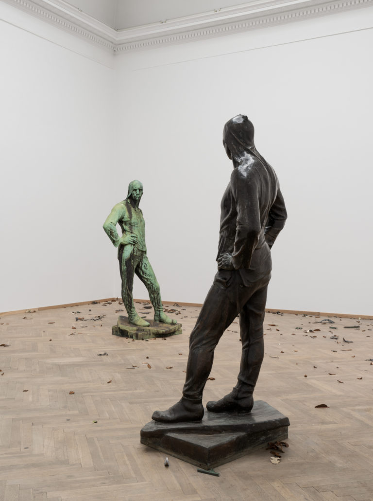 A bright exhibition room with two bronze sculptures, one more weathered than the other, standing opposite each other. The sculptures are almost identical, depicting a male human body draped in cloth. The figures are looking directly at each other, with one hand resting on their hips. Small leaves of bronze are spread out on the floor.