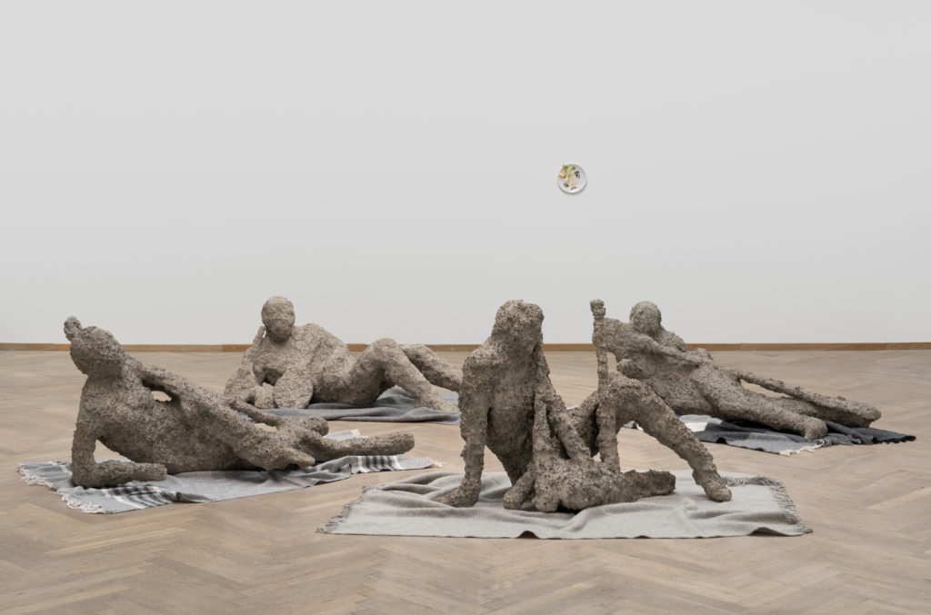 Four sculptures are lying on the floor, on carpets in an exhibition room.