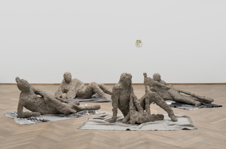 Four sculptures are lying on the floor, on carpets in an exhibition room.