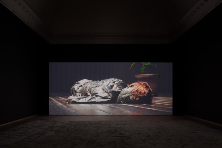 A dark exhibition room with a screen set up at the back wall, displaying an animated video work of a sunken human body lying on a wooden floor with dark leather looking skin and red hair. The animation is reminiscent of the Danish bog body Grauballe Man.