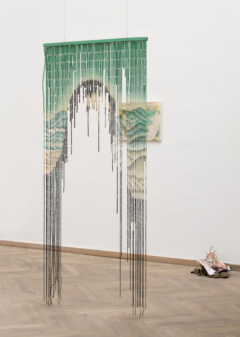 I et lyst udstillingsrum, hvor et dørgardin hænger ned fra loftet. Gardinet er klippet op i midten, og et motiv af en kvinde kan anes.