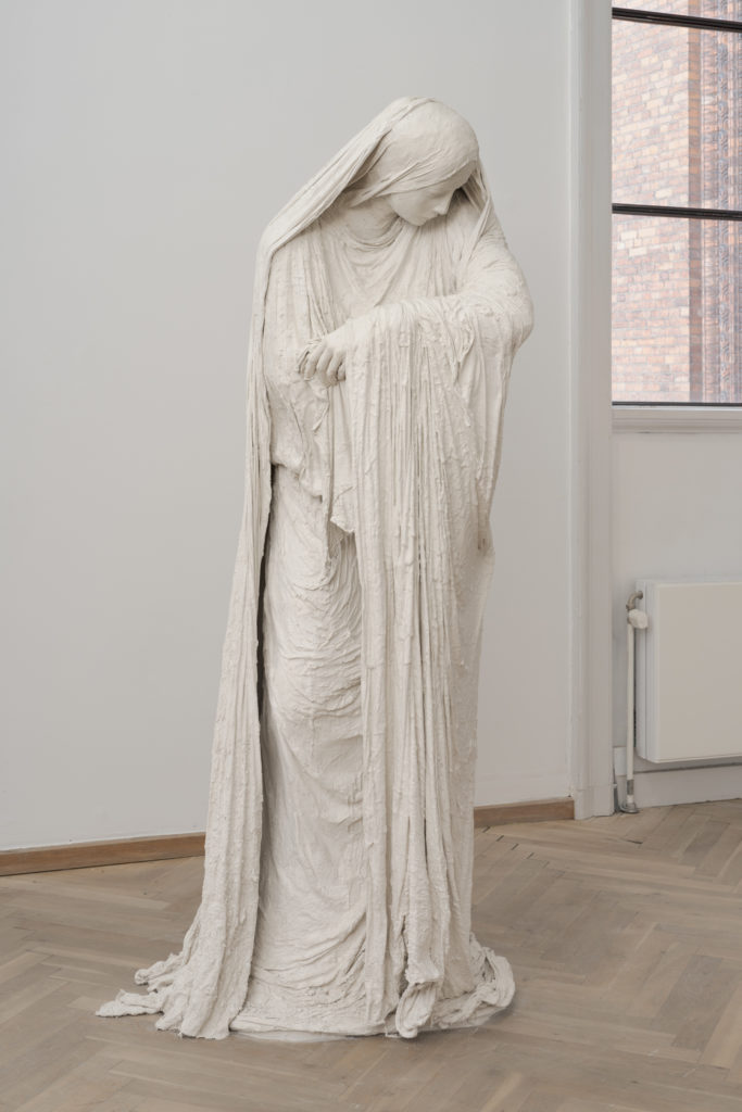 En skulptur af en kvinde iklædt klæder på krop og hovede. Kvinden kigger ned mod sine arme, som er tomme.