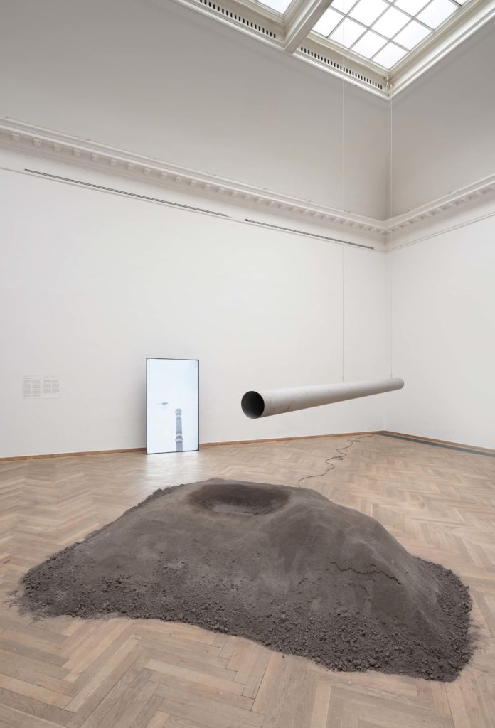 In the center of an exhibition space lies a pile of coal, with a pipe hanging above it.
