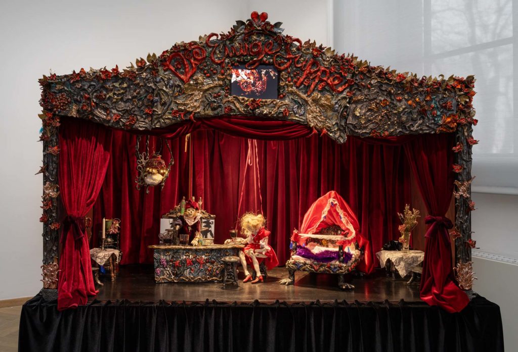 A puppet theater with puppets and miniature furniture, surrounded by red velvet.