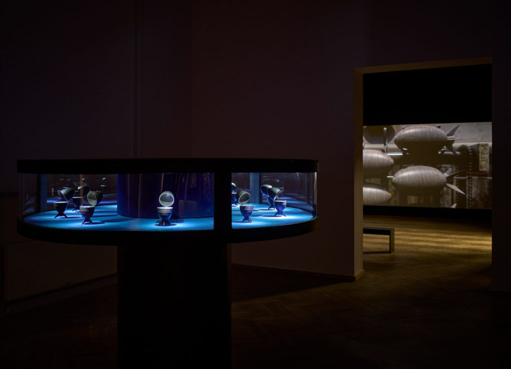 A glass display case in an exhibition space; in the background, a film can be glimpsed, projected onto the wall.