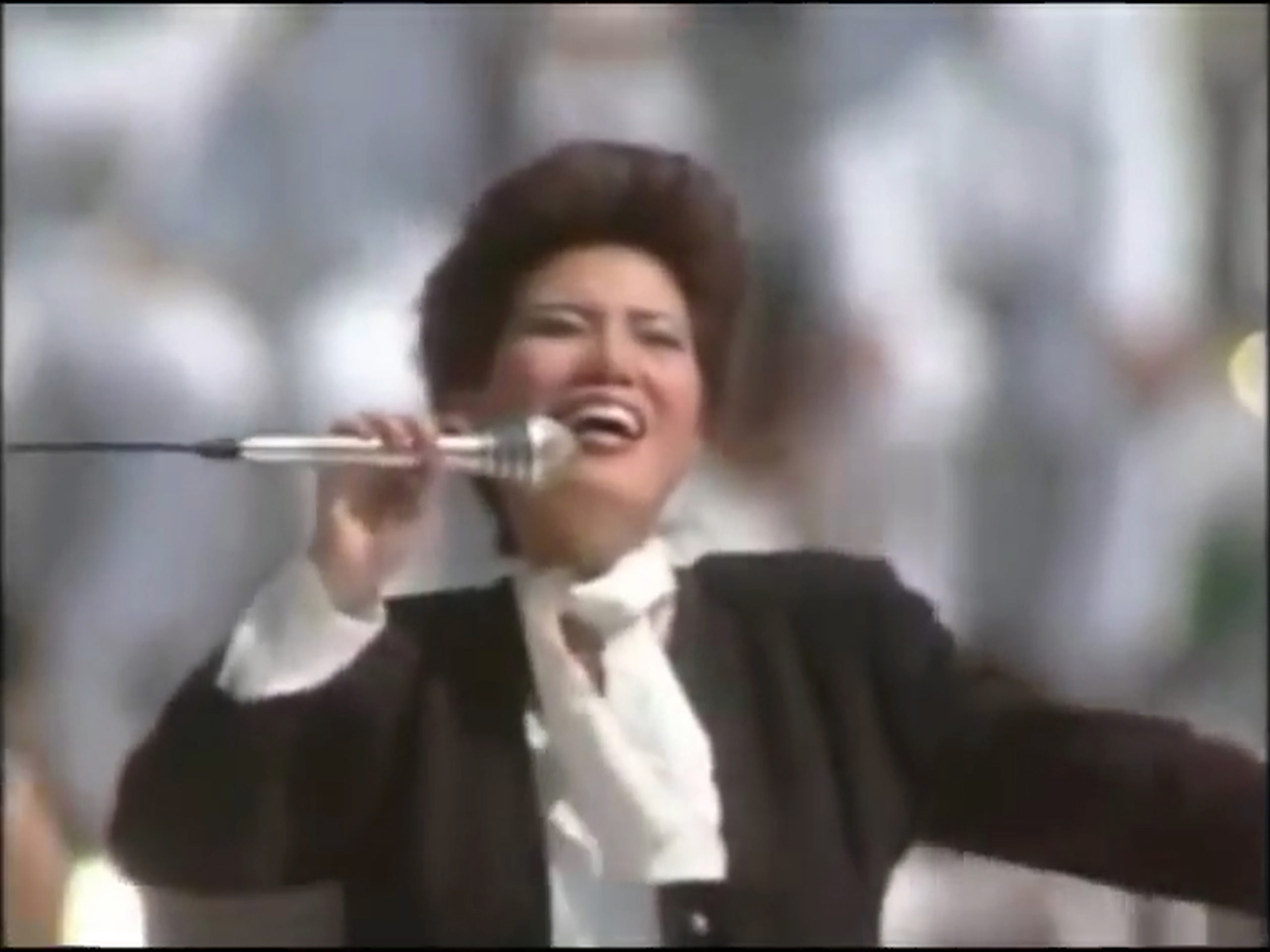 Kvinde med kort hår synger energisk i mikrofon iført sort jakke og hvid skjorte med sløjfe, foran en sløret baggrund af folk i hvidt.